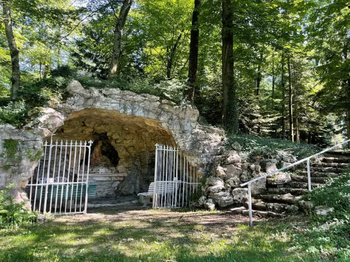 La grotte Sainte-Madeleine, chapelle naturelle en pierre au cœur du bois, avec grille blanche et escaliers en pierre à droite.
